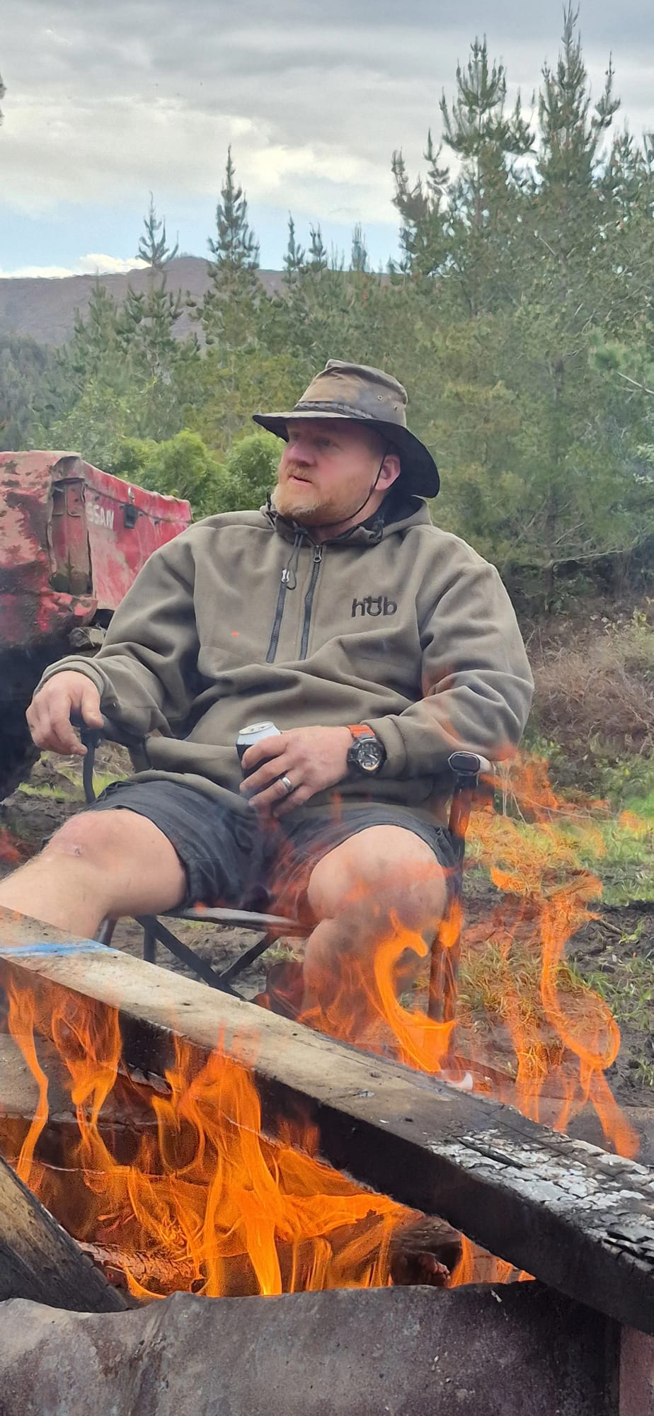 Kiwi adventurer in Full Send Hub Fleece Collection hoodie by campfire on a New Zealand bush trail, windproof and water-resistant for off-road camping.
