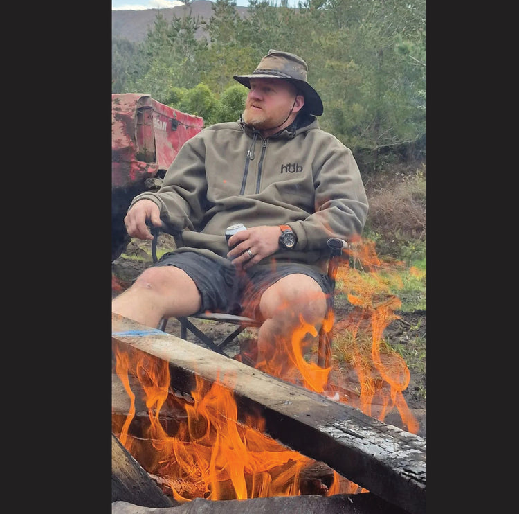 Kiwi adventurer in Full Send Hub Fleece Collection hoodie by campfire on a New Zealand bush trail, windproof and water-resistant for off-road camping.